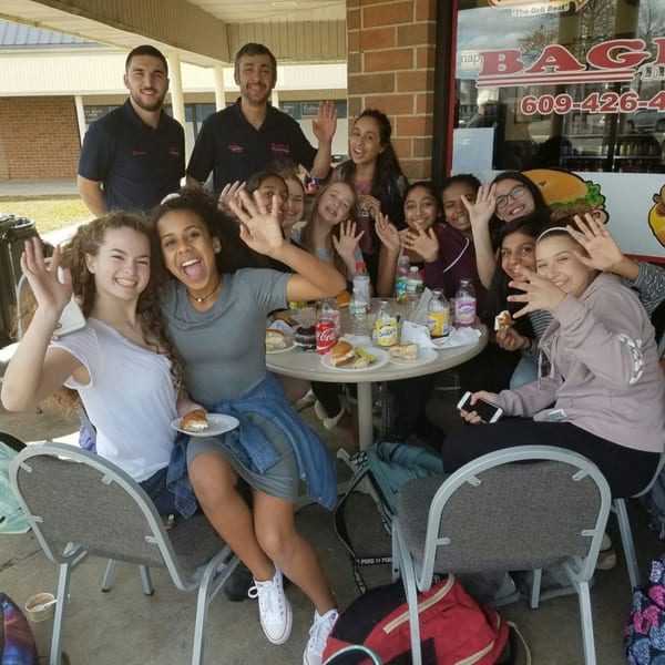 Two Bagels N' Cream staff members and a group of young women wave outside the bagel shop.