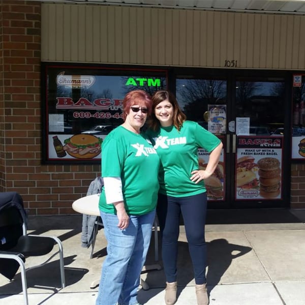 Two women in green shirts stand smiling outside Bagels N' Cream, a bagel shop.