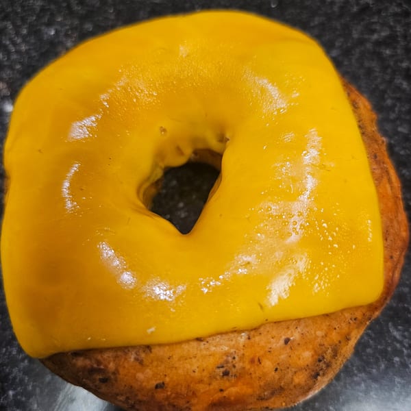 A Bagels N' Cream bagel topped with melted yellow cheese, on a dark surface.