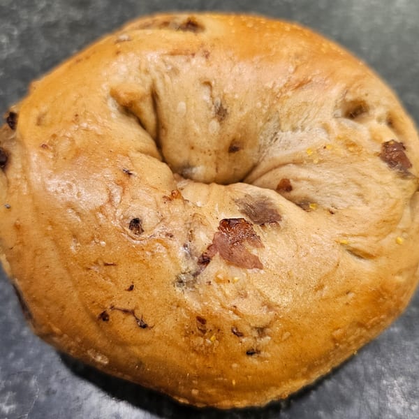 A golden-brown bagel with visible specks of dried fruit and spices on a dark surface.