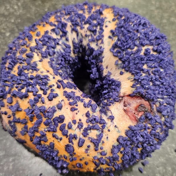 A Bagels N' Cream bagel, generously coated with vibrant purple sprinkles, on a dark surface.