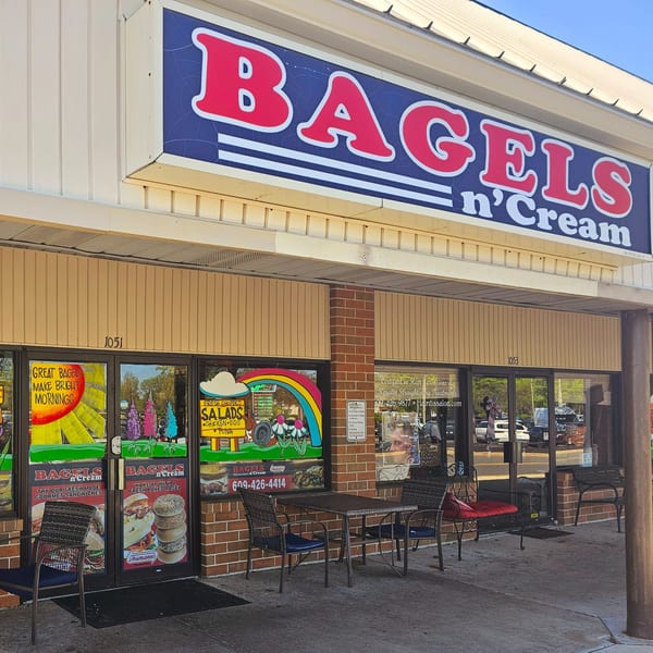 Exterior of Bagels N' Cream with a blue and red sign, outdoor seating, and window advertisements.