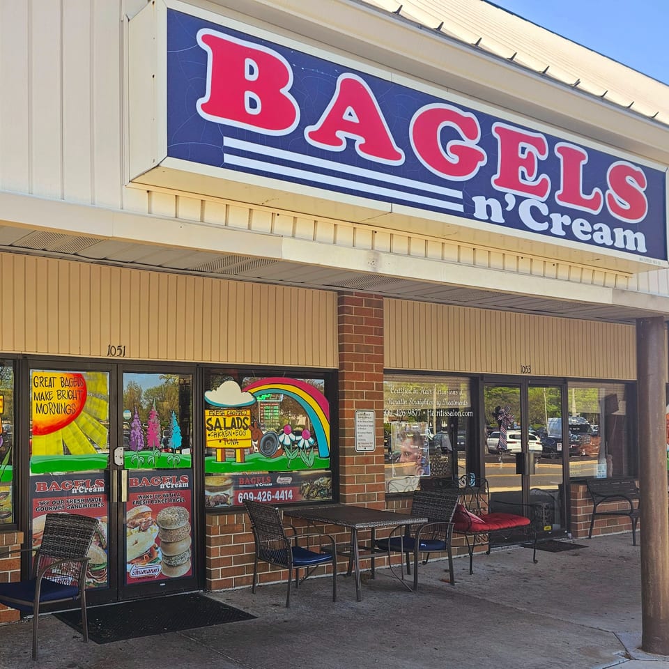 Exterior of Bagels N' Cream with a blue and red sign, outdoor seating, and window advertisements.