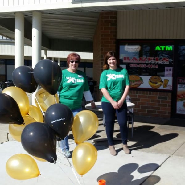 Two women in green shirts stand outside Bagels N' Cream, with black and gold balloons in the foreground.
