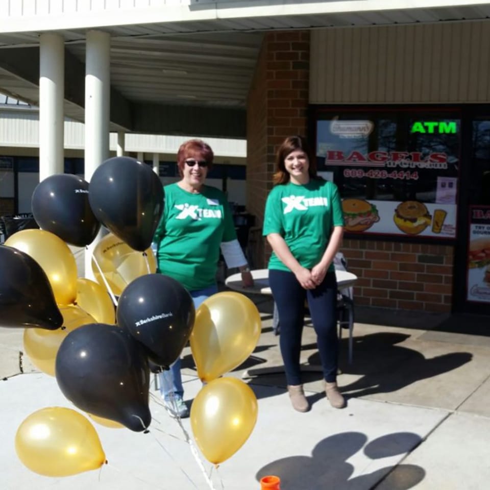 Two women in green shirts stand outside Bagels N' Cream, with black and gold balloons in the foreground.