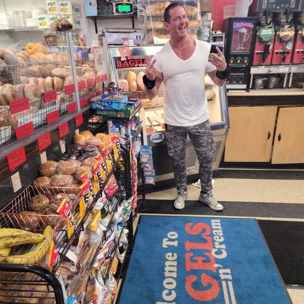 A man smiles in Bagels N' Cream, surrounded by shelves of bagels, muffins, and snacks.