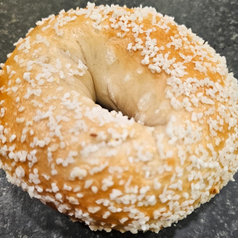 A close-up of a golden-brown bagel from Bagels N' Cream, generously topped with coarse salt.