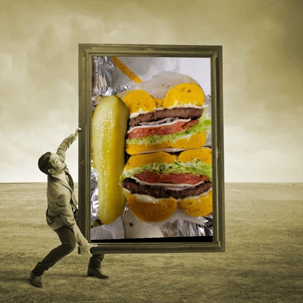 A man holds a framed picture of two bagel burgers with lettuce, tomato, and a pickle.