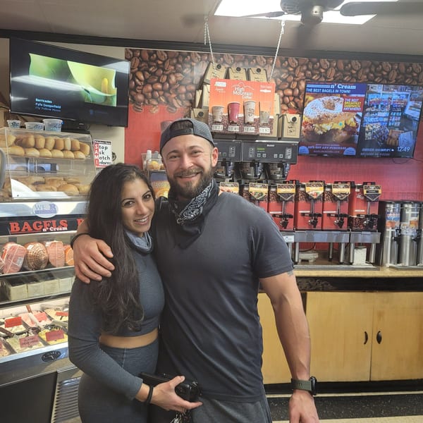A smiling couple stands in front of Bagels N' Cream counter with bagels, coffee, and cream cheese.