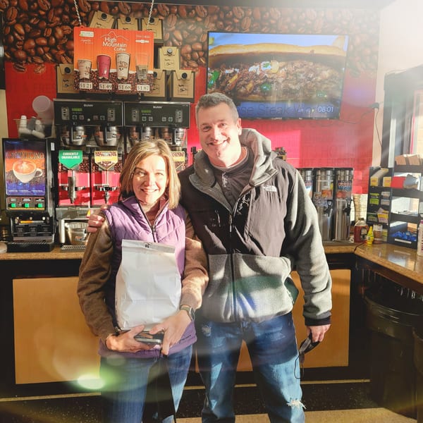 Two smiling customers stand inside Bagels N' Cream, holding a white bag near coffee machines.