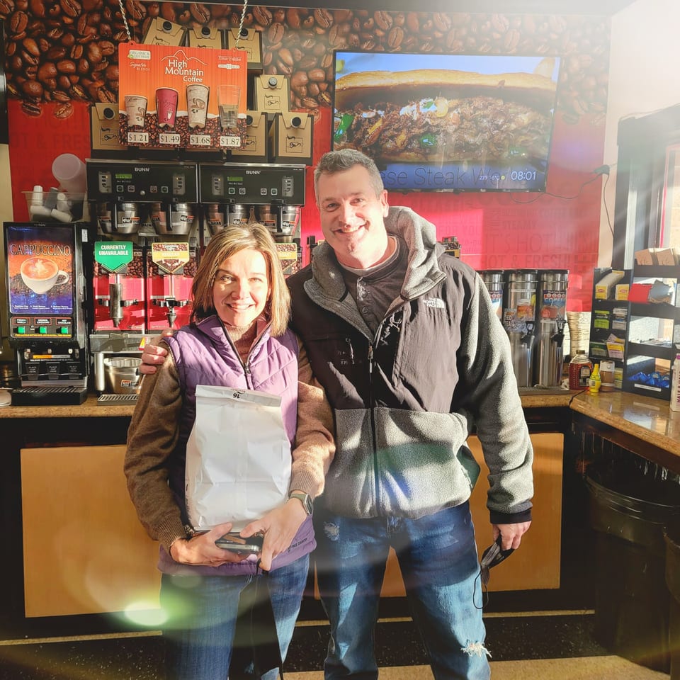 Two smiling customers stand inside Bagels N' Cream, holding a white bag near coffee machines.