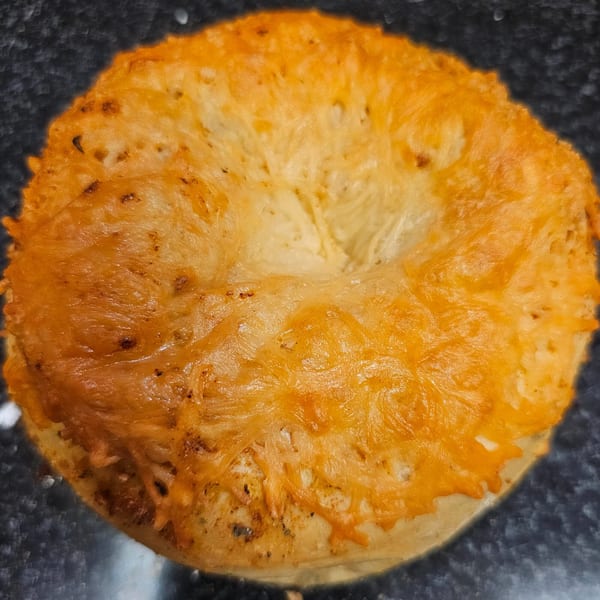 A golden-brown cheese bagel from Bagels N' Cream, topped with melted cheddar, on a dark surface.