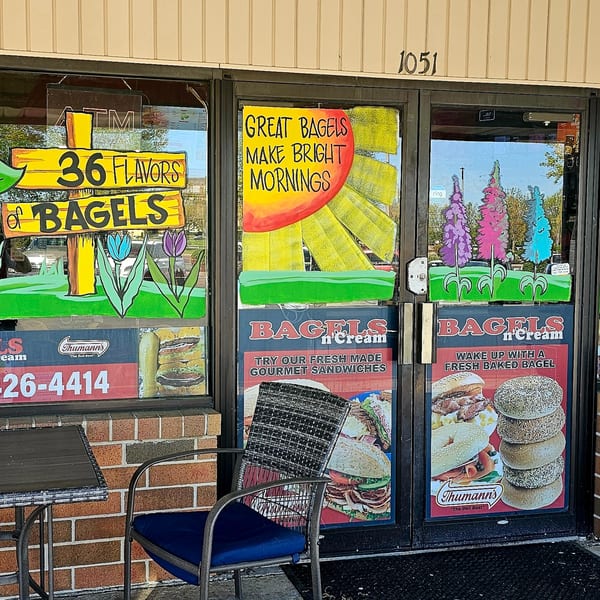 The Bagels N' Cream storefront features colorful window art, advertisements for bagels and sandwiches, and outdoor seating.