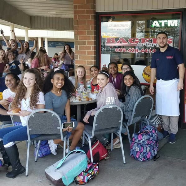 Smiling customers and a staff member outside Bagels N' Cream, with bagels and drinks on tables.