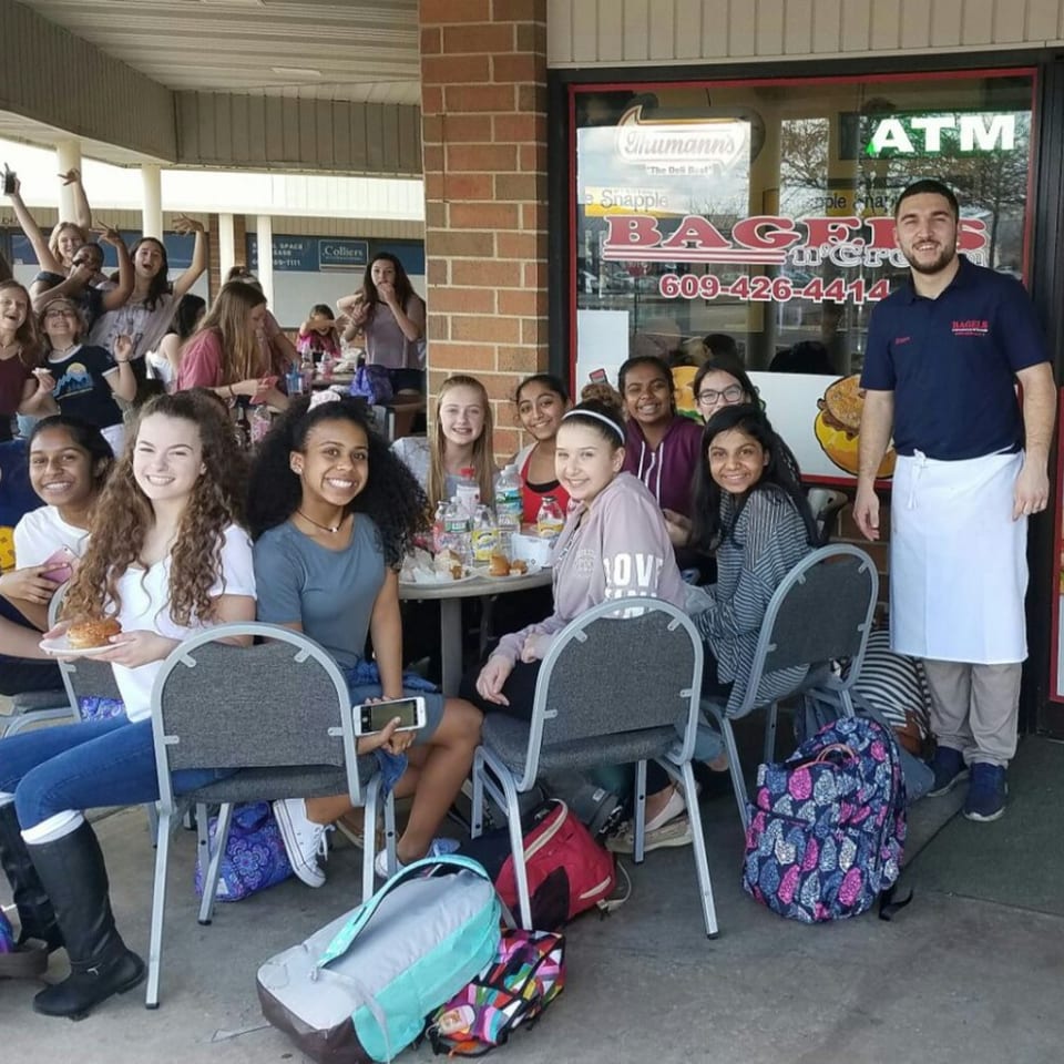 Smiling customers and a staff member outside Bagels N' Cream, with bagels and drinks on tables.