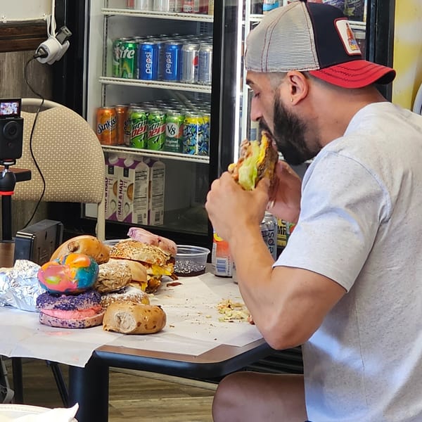 Man in a cap eating a large bagel sandwich at Bagels N' Cream, with other bagels on the table.