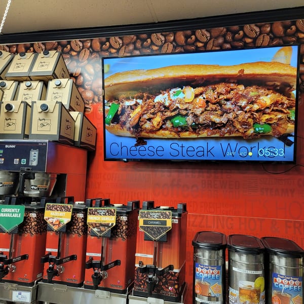 A coffee station with various flavored coffee dispensers and a screen displaying a cheesesteak.